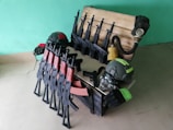 A collection of replica rifles and handguns is arranged neatly in and around a wooden crate, accompanied by helmets and a gas mask. The weapons have distinct black bodies with brown and red accents. The gas mask is attached to a hose leading into the crate, and the helmets are dark gray with neon green detailing. The background features a plain teal wall.