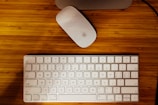 A white wireless keyboard and an Apple Magic Mouse are placed on a wooden table. The keyboard has English and Chinese characters on its keys.