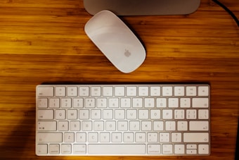 A white wireless keyboard and an Apple Magic Mouse are placed on a wooden table. The keyboard has English and Chinese characters on its keys.