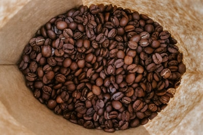 A close-up view of roasted coffee beans inside a brown paper bag. The beans are dark and glossy with various shades of brown, highlighting their rich texture.