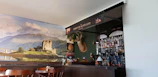 A cozy pub interior featuring a bar with an array of bottles and a decorative backdrop. The wall behind the bar displays a large scenic photo of a castle on a loch, with lush green hills and a partly cloudy sky. Mounted above the bar is a deer head with antlers, and various items like a Scottish flag and other decorations contribute to a Scottish theme.