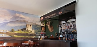 A cozy pub interior featuring a bar with an array of bottles and a decorative backdrop. The wall behind the bar displays a large scenic photo of a castle on a loch, with lush green hills and a partly cloudy sky. Mounted above the bar is a deer head with antlers, and various items like a Scottish flag and other decorations contribute to a Scottish theme.