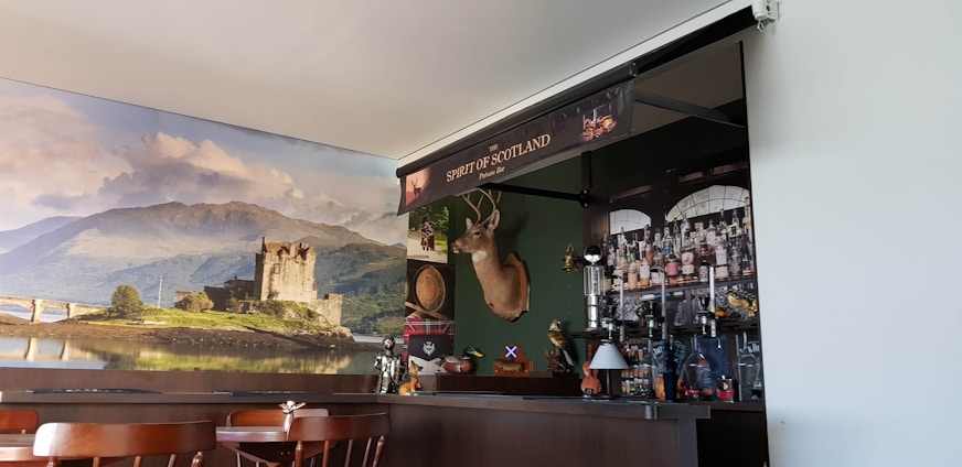 A cozy pub interior featuring a bar with an array of bottles and a decorative backdrop. The wall behind the bar displays a large scenic photo of a castle on a loch, with lush green hills and a partly cloudy sky. Mounted above the bar is a deer head with antlers, and various items like a Scottish flag and other decorations contribute to a Scottish theme.