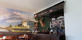 A cozy pub interior featuring a bar with an array of bottles and a decorative backdrop. The wall behind the bar displays a large scenic photo of a castle on a loch, with lush green hills and a partly cloudy sky. Mounted above the bar is a deer head with antlers, and various items like a Scottish flag and other decorations contribute to a Scottish theme.
