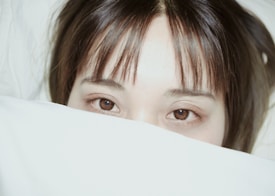 A person's eyes peek out from under a white blanket or cover. The individual has dark hair and brown eyes, and the image captures only the upper part of their face, creating an intimate and serene atmosphere.