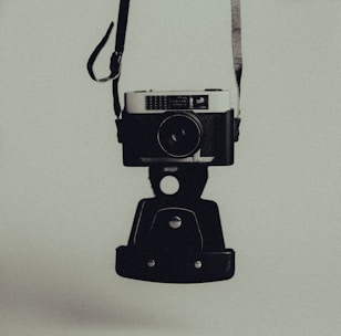 A vintage camera is suspended by its strap against a muted background. The camera has a classic design with a prominent lens and metal body. The overall image has a dark and moody aesthetic.