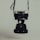 A vintage camera is suspended by its strap against a muted background. The camera has a classic design with a prominent lens and metal body. The overall image has a dark and moody aesthetic.
