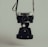 A vintage camera is suspended by its strap against a muted background. The camera has a classic design with a prominent lens and metal body. The overall image has a dark and moody aesthetic.