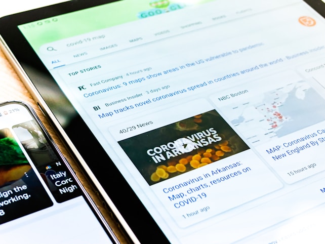 A close-up view of two electronic devices displaying news articles related to COVID-19. The tablet screen shows a news website with headlines about coronavirus charts, maps, and resources. The phone screen on the left partially shows more COVID-19 related content with a focus on Italy.