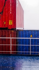 A stack of colorful shipping containers in red and blue is seen on a wet surface on a rainy day. The containers have caution signs and numbers printed on them. The ground is reflective due to the rain.