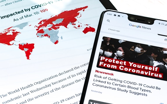 A smartphone displays a news alert about COVID-19, with a headline urging people to protect themselves from the virus. The screen shows a group of people wearing masks, emphasizing health precautions. In the background, a world map highlights countries impacted by COVID-19 in red, and text from a report or article is partially visible.