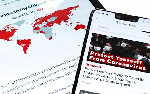 A smartphone displays a news alert about COVID-19, with a headline urging people to protect themselves from the virus. The screen shows a group of people wearing masks, emphasizing health precautions. In the background, a world map highlights countries impacted by COVID-19 in red, and text from a report or article is partially visible.