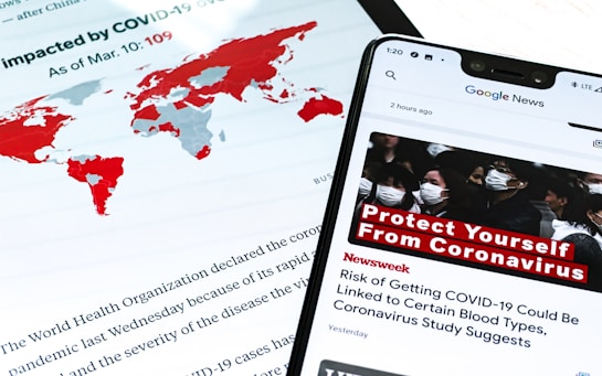 A smartphone displays a news alert about COVID-19, with a headline urging people to protect themselves from the virus. The screen shows a group of people wearing masks, emphasizing health precautions. In the background, a world map highlights countries impacted by COVID-19 in red, and text from a report or article is partially visible.