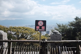 A no smoking sign is positioned against a scenic backdrop of a city with trees and mountains. The sign includes both English and Japanese text, surrounded by a wooden fence and stone structures in the foreground. The sky is partially cloudy.