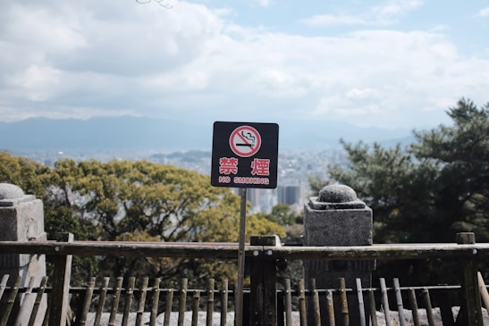A no smoking sign is positioned against a scenic backdrop of a city with trees and mountains. The sign includes both English and Japanese text, surrounded by a wooden fence and stone structures in the foreground. The sky is partially cloudy.