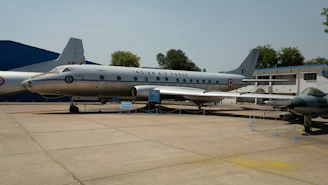 A silver aircraft with markings of the Indian Air Force is displayed outdoors on a concrete surface, accompanied by informational signs. The aircraft is surrounded by ropes and poles, with another smaller aircraft visible to the right. Trees and a building are seen in the background under clear skies.