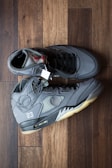 A pair of dark gray high-top sneakers with black laces, displayed on a wooden floor. The shoes have a visible air unit in the midsole, various stitched panels, and a number 23 near the heel. The lining and tongue feature different shades of gray, with a visible tag attached.