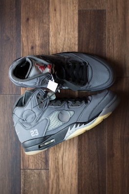 A pair of dark gray high-top sneakers with black laces, displayed on a wooden floor. The shoes have a visible air unit in the midsole, various stitched panels, and a number 23 near the heel. The lining and tongue feature different shades of gray, with a visible tag attached.