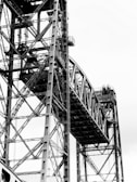 A detailed view of a large steel bridge structure with complex intersecting beams and cables forming a geometric pattern against a light sky.