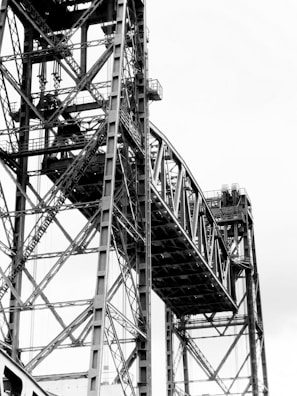 A detailed view of a large steel bridge structure with complex intersecting beams and cables forming a geometric pattern against a light sky.