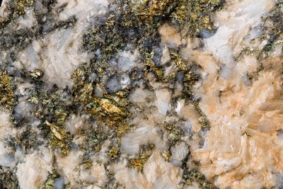 A close-up view of a textured mineral surface featuring a mixture of gold, white, and greyish-blue crystalline structures. The gold elements shimmer amidst the rough, irregular patterns of the surrounding minerals.
