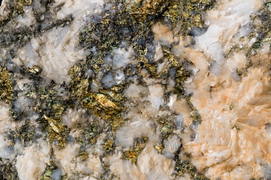 A close-up view of a textured mineral surface featuring a mixture of gold, white, and greyish-blue crystalline structures. The gold elements shimmer amidst the rough, irregular patterns of the surrounding minerals.