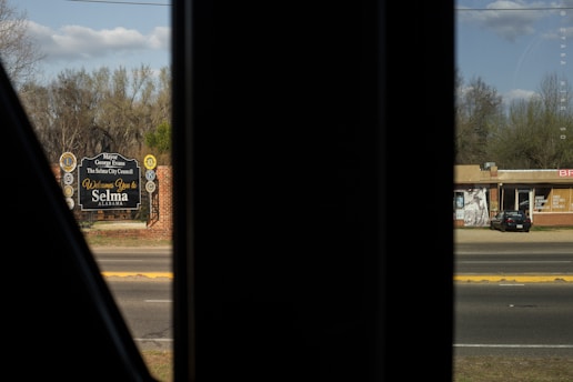 The image captures a street scene with a sign that reads 'Welcome to Selma, Alabama'. The sign is set against a backdrop of trees. In the foreground, there is a dark section likely resembling a window frame. Across the street, there is a parked car in front of a brick building with large windows.