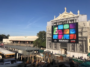 An outdoor setting featuring a large, historical building with a modern billboard advertising the Netflix series 'Maniac.' The billboard displays colorful images of faces segmented into nine parts, with bright blue, green, red, and pink hues. The building facade is ornate, with statues on top. Below the billboard, the area is busy with people walking and boats docked by a canal.