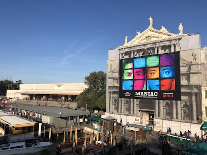 An outdoor setting featuring a large, historical building with a modern billboard advertising the Netflix series 'Maniac.' The billboard displays colorful images of faces segmented into nine parts, with bright blue, green, red, and pink hues. The building facade is ornate, with statues on top. Below the billboard, the area is busy with people walking and boats docked by a canal.