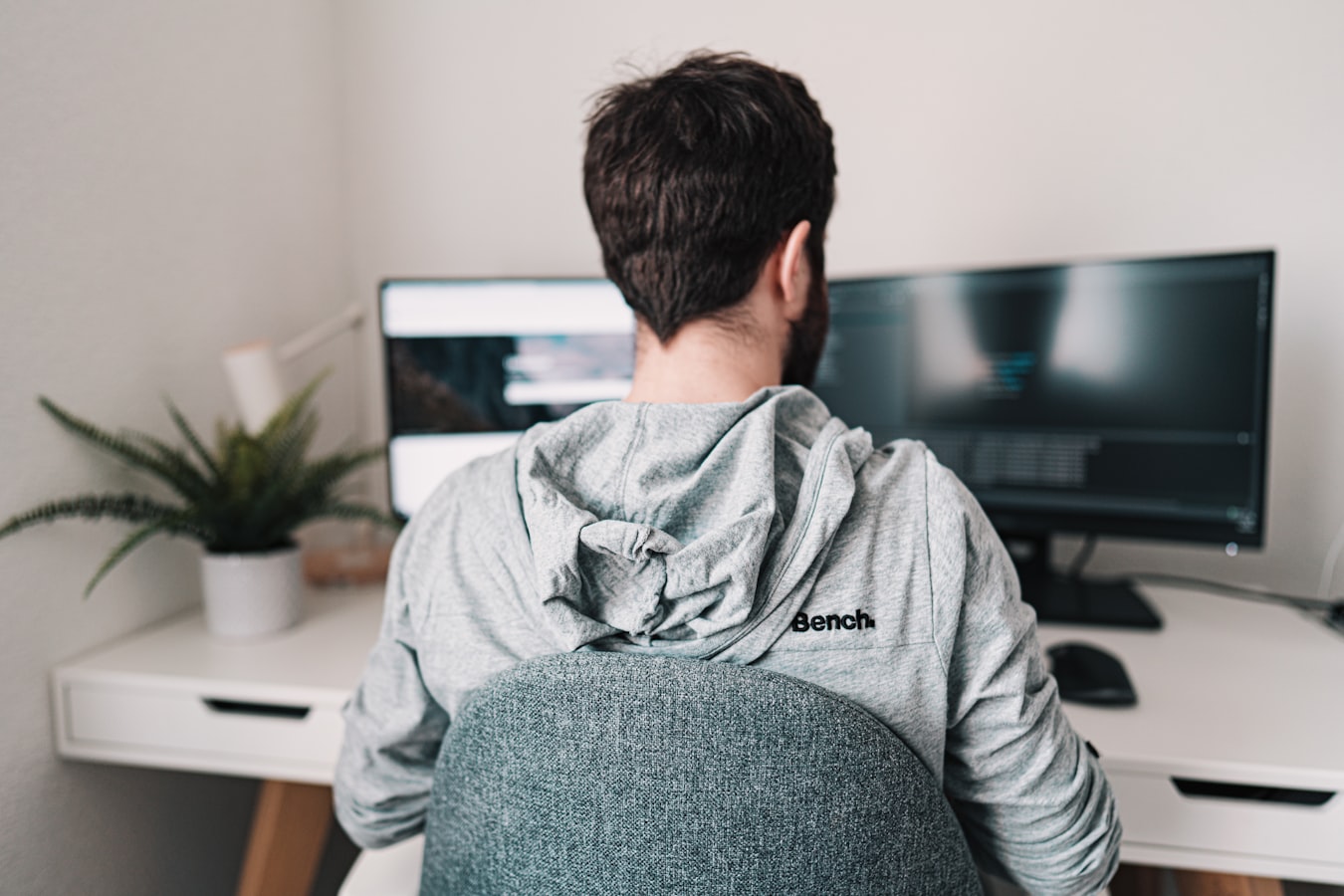 Homem sentado ao computador, em casa, à frente de dois monitores com dados.