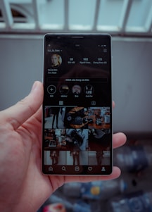 A hand is holding a smartphone displaying a social media profile. The screen shows various images arranged in a grid, along with a profile picture, username, and some numerical details such as posts, followers, and following. The background is blurred, indicating it might be outside.