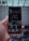 A hand is holding a smartphone displaying a social media profile. The screen shows various images arranged in a grid, along with a profile picture, username, and some numerical details such as posts, followers, and following. The background is blurred, indicating it might be outside.