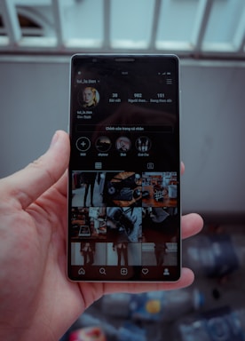 A hand is holding a smartphone displaying a social media profile. The screen shows various images arranged in a grid, along with a profile picture, username, and some numerical details such as posts, followers, and following. The background is blurred, indicating it might be outside.