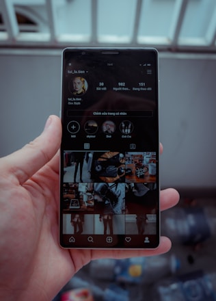 A hand is holding a smartphone displaying a social media profile. The screen shows various images arranged in a grid, along with a profile picture, username, and some numerical details such as posts, followers, and following. The background is blurred, indicating it might be outside.