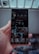 A hand is holding a smartphone displaying a social media profile. The screen shows various images arranged in a grid, along with a profile picture, username, and some numerical details such as posts, followers, and following. The background is blurred, indicating it might be outside.