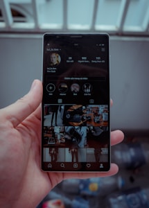 A hand is holding a smartphone displaying a social media profile. The screen shows various images arranged in a grid, along with a profile picture, username, and some numerical details such as posts, followers, and following. The background is blurred, indicating it might be outside.
