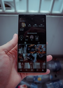 A hand is holding a smartphone displaying a social media profile. The screen shows various images arranged in a grid, along with a profile picture, username, and some numerical details such as posts, followers, and following. The background is blurred, indicating it might be outside.