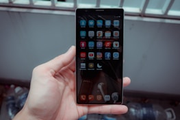 A hand holds a smartphone with a grid of app icons displayed on the screen. The apps include popular social media platforms and tools, arranged neatly. The background is blurred, consisting of bottles and a metal fence.