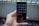 A hand holds a smartphone with a grid of app icons displayed on the screen. The apps include popular social media platforms and tools, arranged neatly. The background is blurred, consisting of bottles and a metal fence.