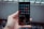 A hand holds a smartphone with a grid of app icons displayed on the screen. The apps include popular social media platforms and tools, arranged neatly. The background is blurred, consisting of bottles and a metal fence.