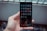 A hand holds a smartphone with a grid of app icons displayed on the screen. The apps include popular social media platforms and tools, arranged neatly. The background is blurred, consisting of bottles and a metal fence.