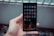 A hand holds a smartphone with a grid of app icons displayed on the screen. The apps include popular social media platforms and tools, arranged neatly. The background is blurred, consisting of bottles and a metal fence.