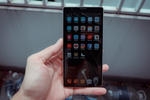 A hand holds a smartphone with a grid of app icons displayed on the screen. The apps include popular social media platforms and tools, arranged neatly. The background is blurred, consisting of bottles and a metal fence.