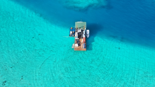 Aerial view of a floating platform on clear turquoise water. The platform seems to be equipped with various items and appears to be a boat or floating structure. The surrounding water transitions from a vibrant light turquoise to a deeper blue.