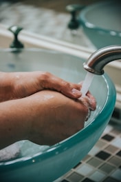 Hands are positioned under running water from a faucet, with a focus on washing and hygiene. The scene includes a glass sink and a tiled surface in the background.