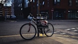 A bicycle is parked on a sidewalk next to a pole in an urban setting. The bike has a sign attached to it displaying advertising material featuring bold text and a red graphic resembling a pointing hand. The background includes a street corner with some buildings, one of which is brick, and there are a few parked cars along the road.