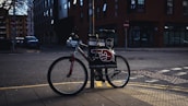 A bicycle is parked on a sidewalk next to a pole in an urban setting. The bike has a sign attached to it displaying advertising material featuring bold text and a red graphic resembling a pointing hand. The background includes a street corner with some buildings, one of which is brick, and there are a few parked cars along the road.