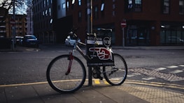 A bicycle is parked on a sidewalk next to a pole in an urban setting. The bike has a sign attached to it displaying advertising material featuring bold text and a red graphic resembling a pointing hand. The background includes a street corner with some buildings, one of which is brick, and there are a few parked cars along the road.