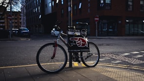 A bicycle is parked on a sidewalk next to a pole in an urban setting. The bike has a sign attached to it displaying advertising material featuring bold text and a red graphic resembling a pointing hand. The background includes a street corner with some buildings, one of which is brick, and there are a few parked cars along the road.
