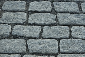 A close-up view of a cobblestone pavement with rectangular stones arranged in a neat, repeating pattern. The surface is slightly rough with visible granules and crevices between the stones.